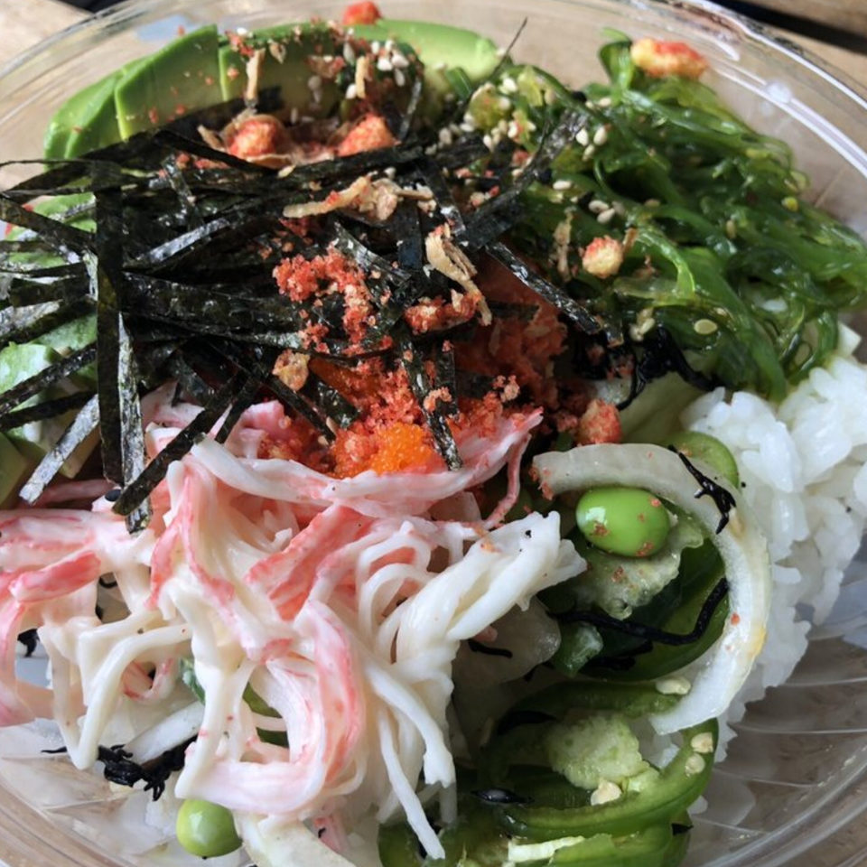Poke bowl with avocado, seaweed, crab, rice, edamame, and jalapeños in a clear container.